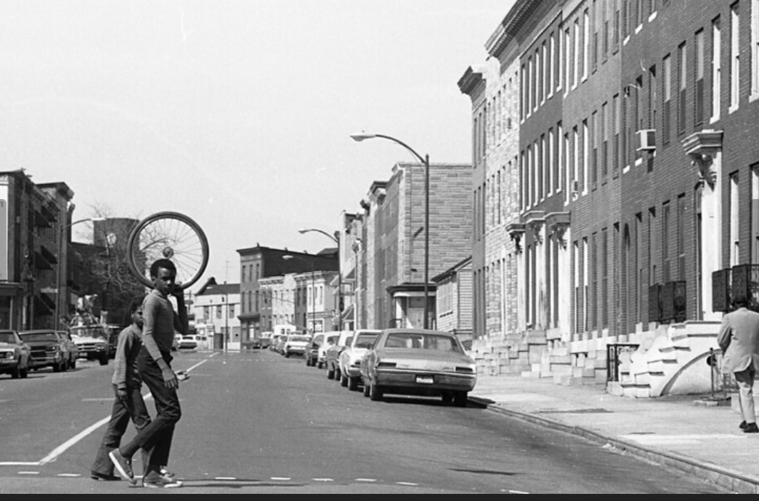 Two young men carry a bicylce wheel across the street.